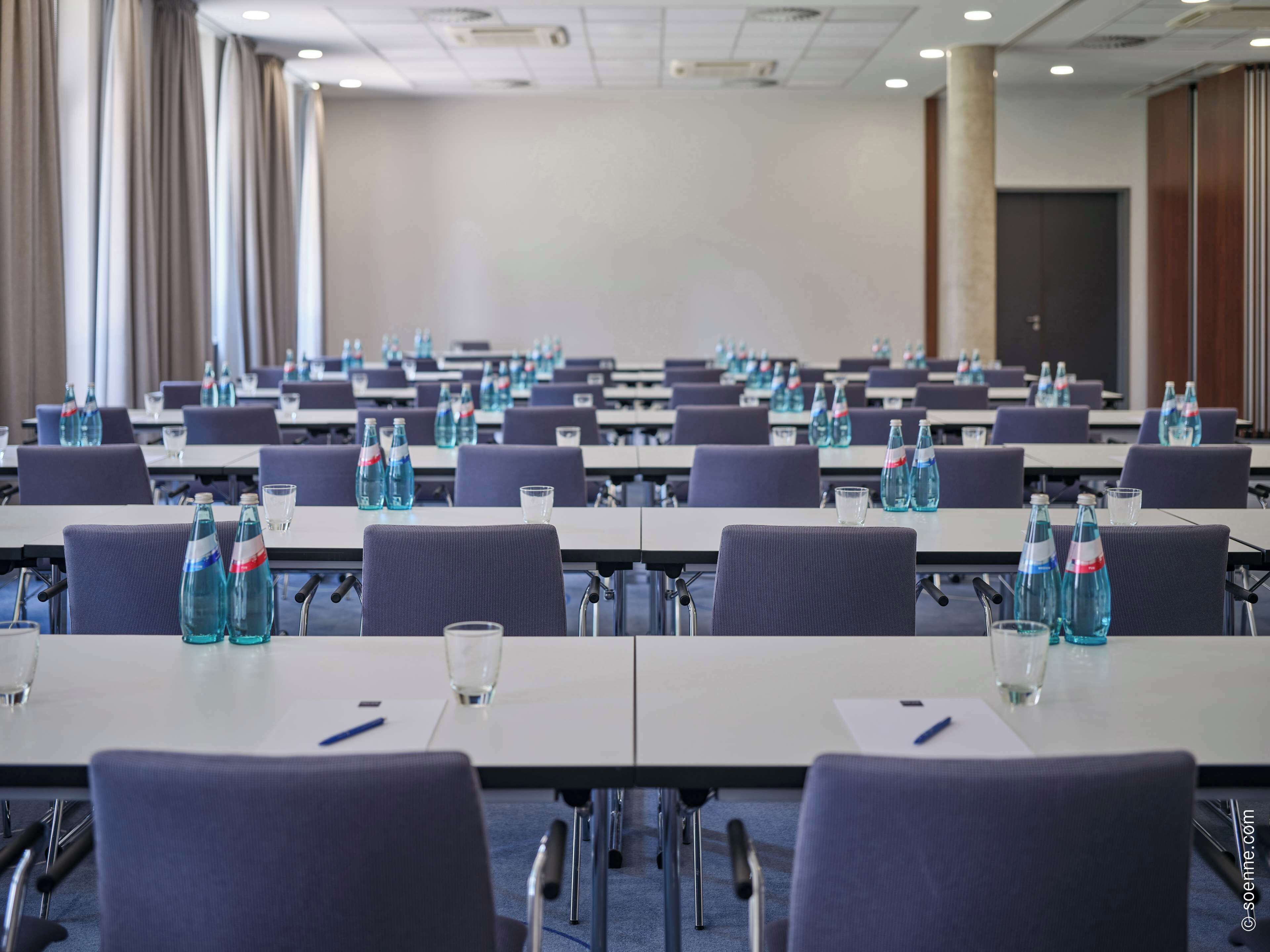 Un salón de conferencias con filas de mesas, sillas y botellas de agua.