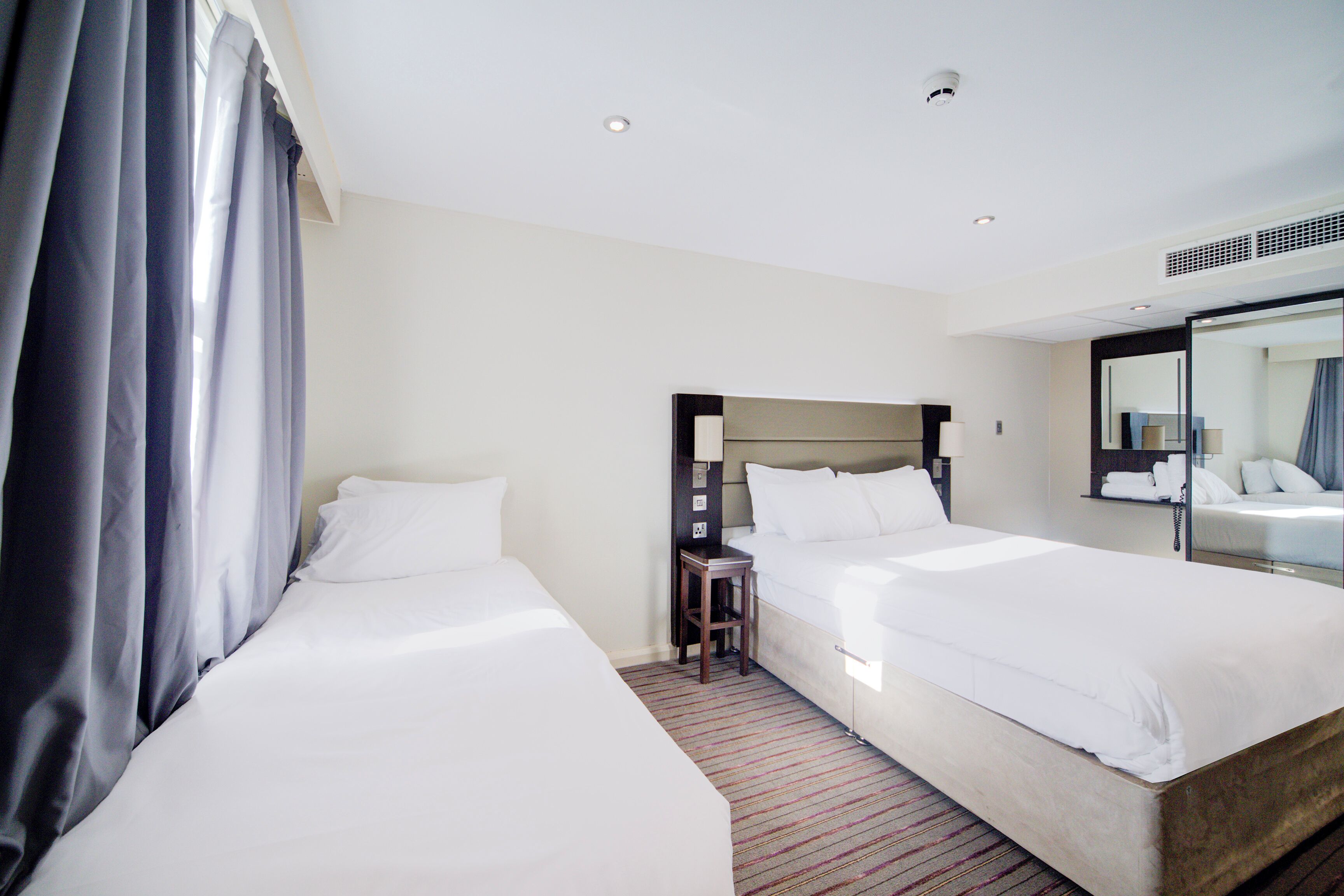 Une chambre d’hôtel avec deux lits, une table de chevet avec une lampe, un miroir et une fenêtre avec des rideaux.