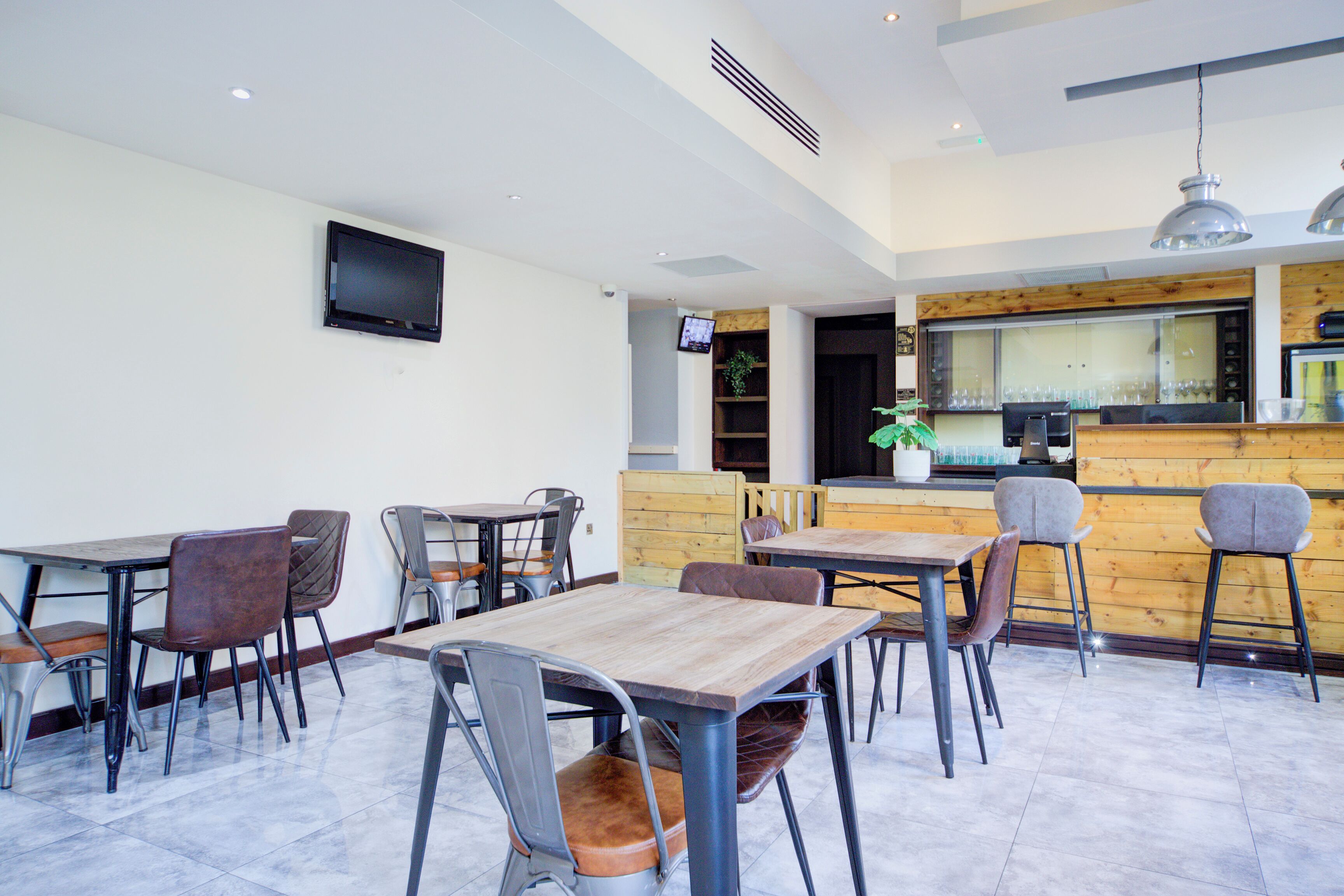 A modern restaurant interior with multiple tables and chairs, a wooden counter, and a flat-screen TV mounted on the wall.