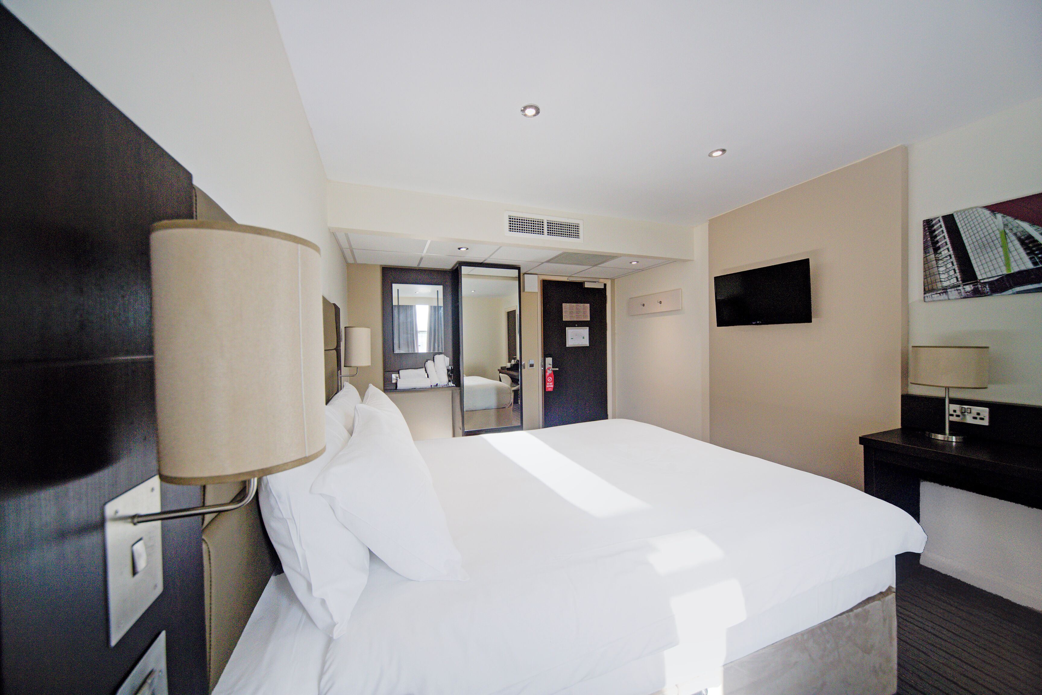 A hotel room with a large bed, a desk with a lamp, a flat-screen TV, and a mirror on the wall.