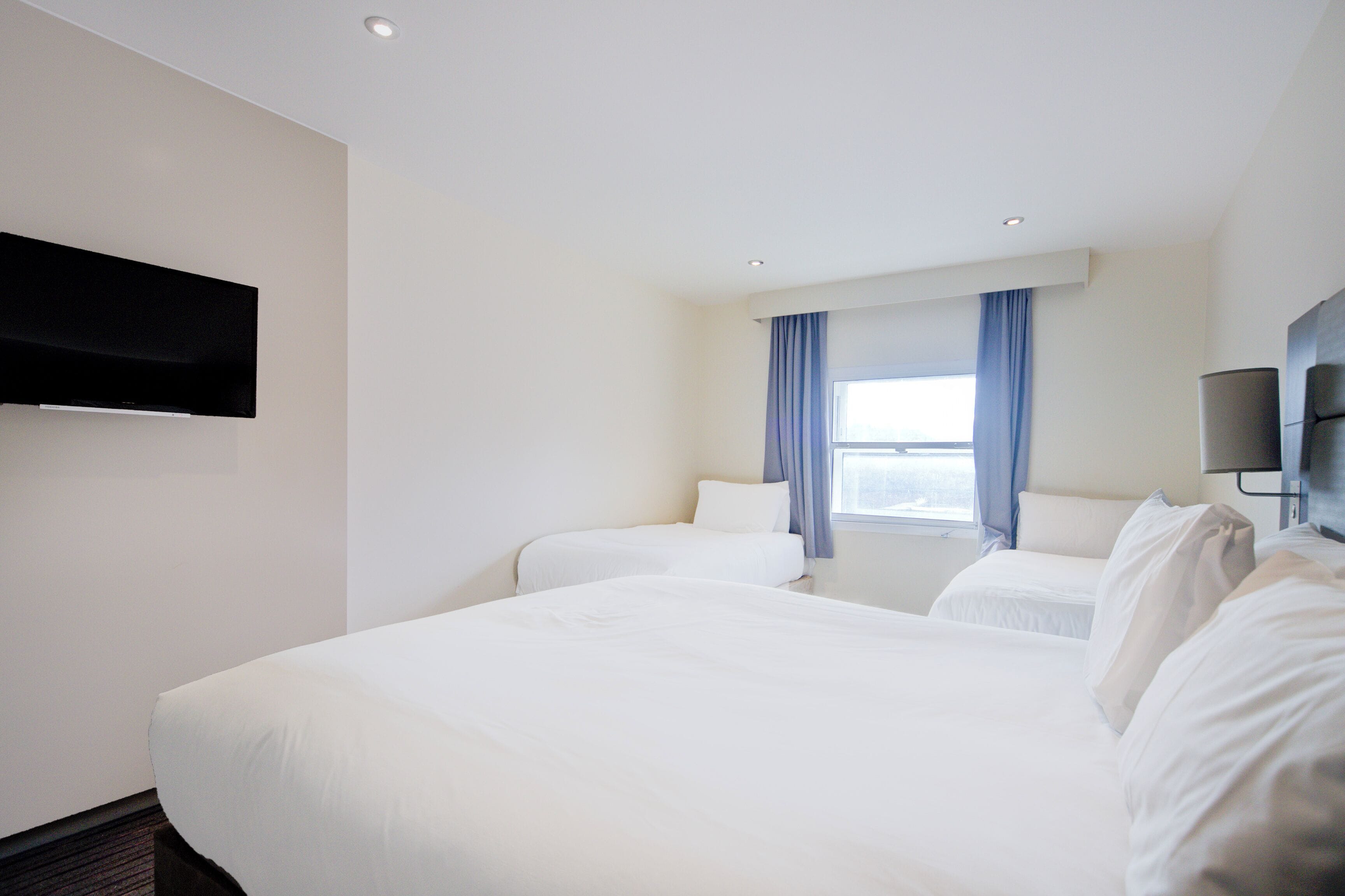 Habitación de hotel con dos camas, un televisor de pantalla plana montado en la pared y una ventana con cortinas azules.