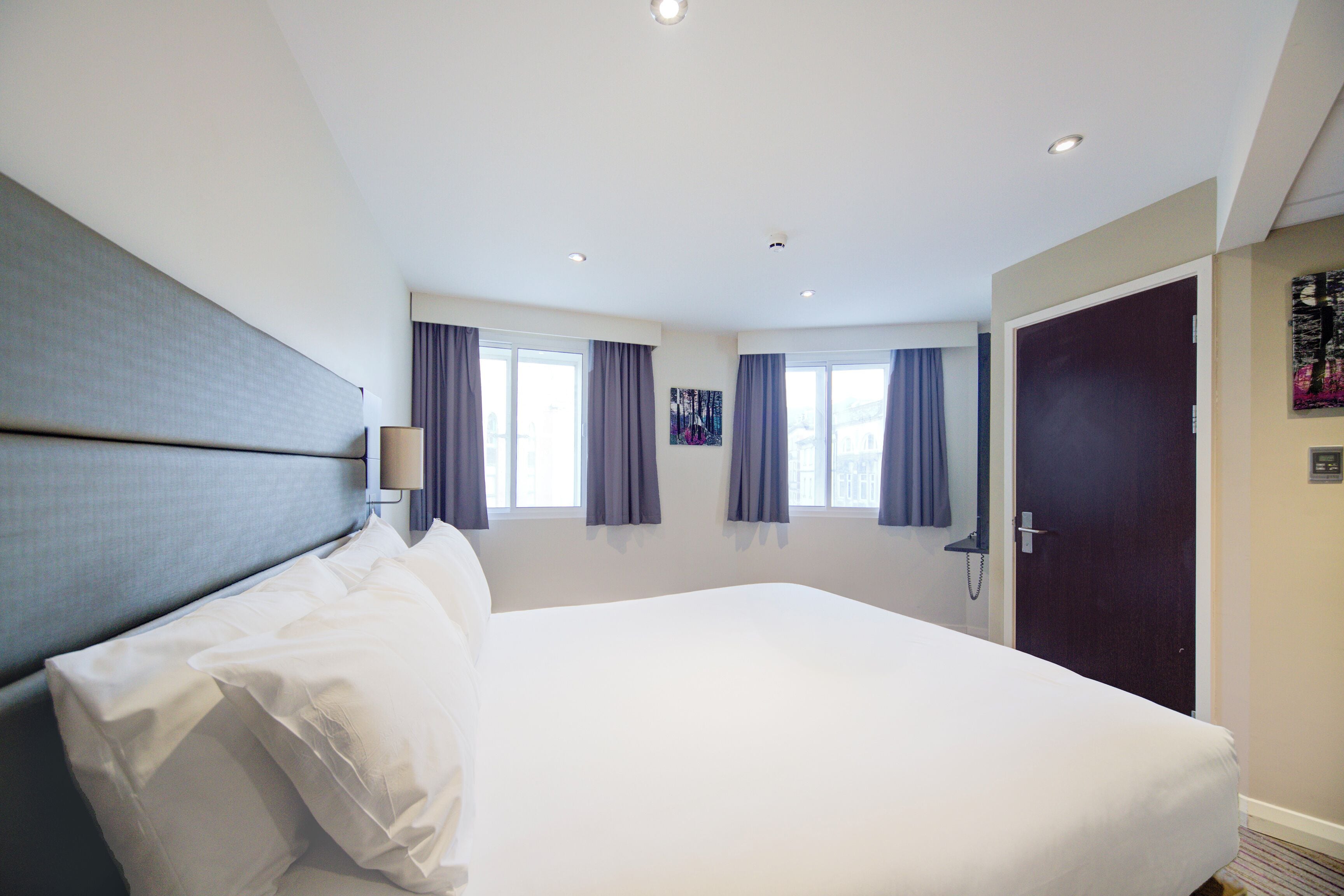 A hotel room with a large bed, two windows with curtains, a door, and a painting on the wall.