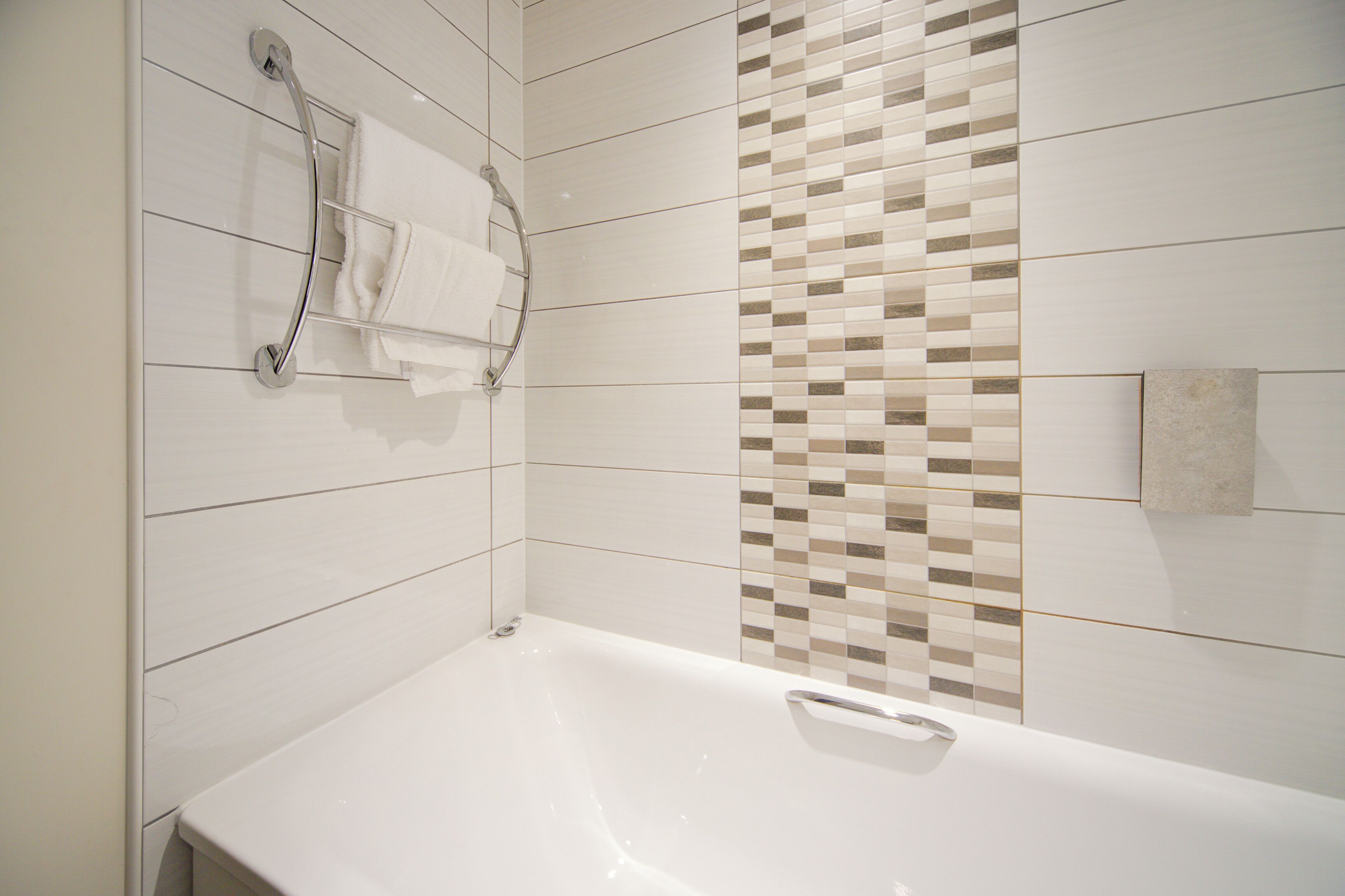 A modern bathroom with a white bathtub, a towel rack, and a mosaic tile wall.