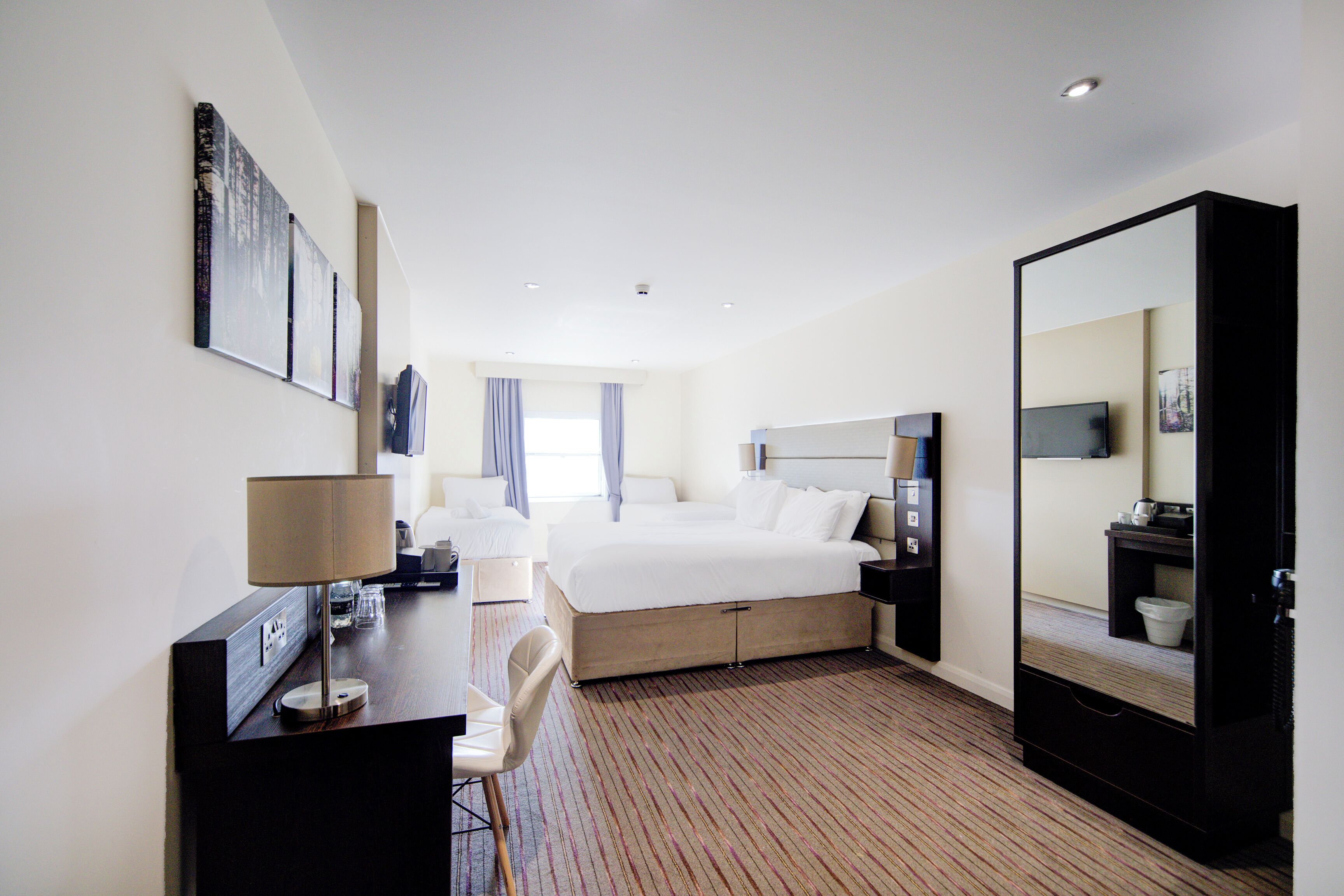 A hotel room with a bed, a desk with a lamp, a chair, a large mirror, and a television.
