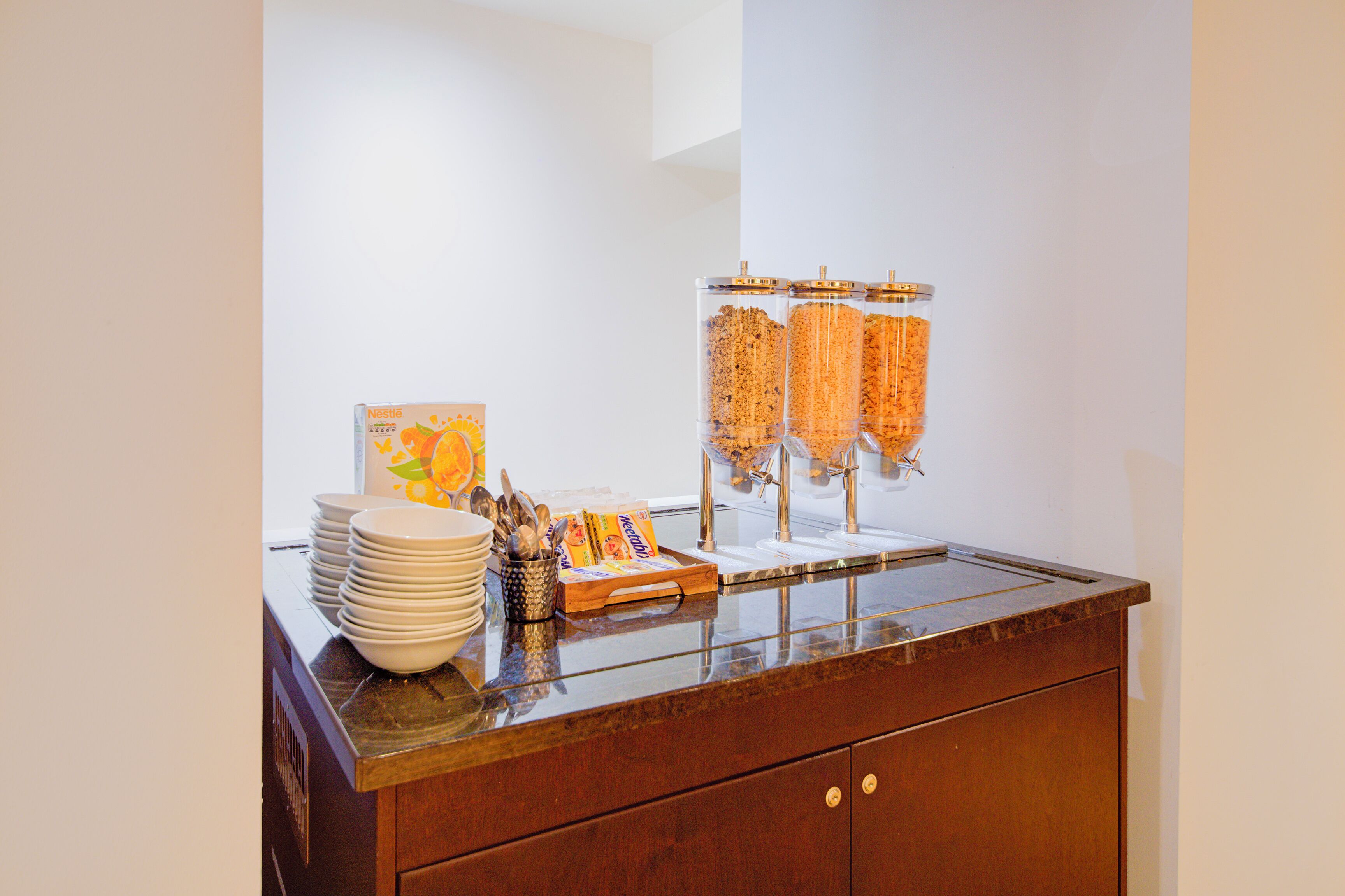 A buffet setup with stacked plates, a tray of utensils, and a cereal dispenser.