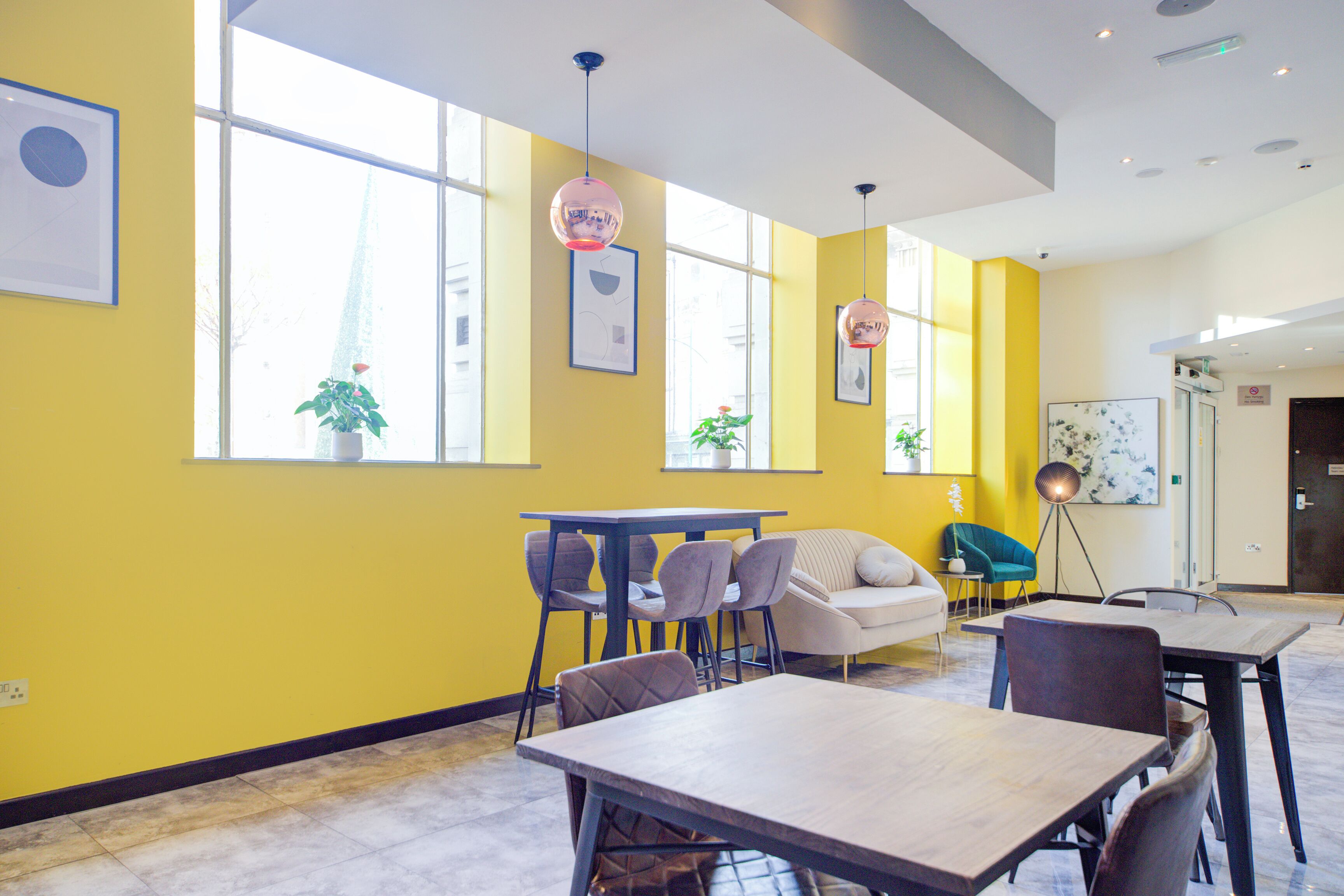 A modern hotel lobby with yellow walls, large windows, a dining area with tables and chairs, and a sofa.