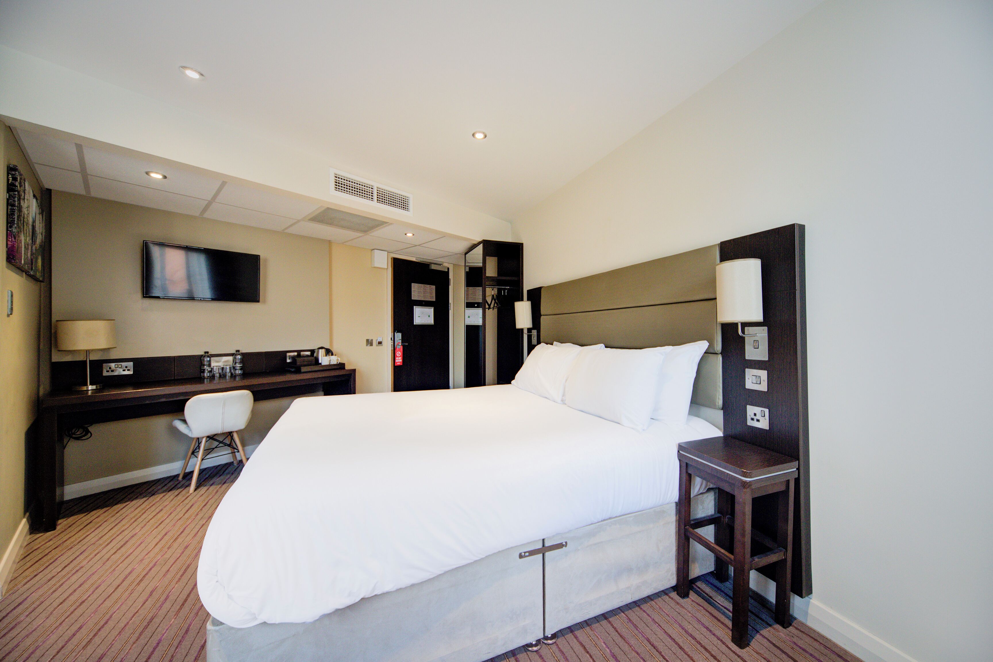 Habitación de hotel con una cama grande, un escritorio, una silla, un televisor y una mesita de noche con lámpara.