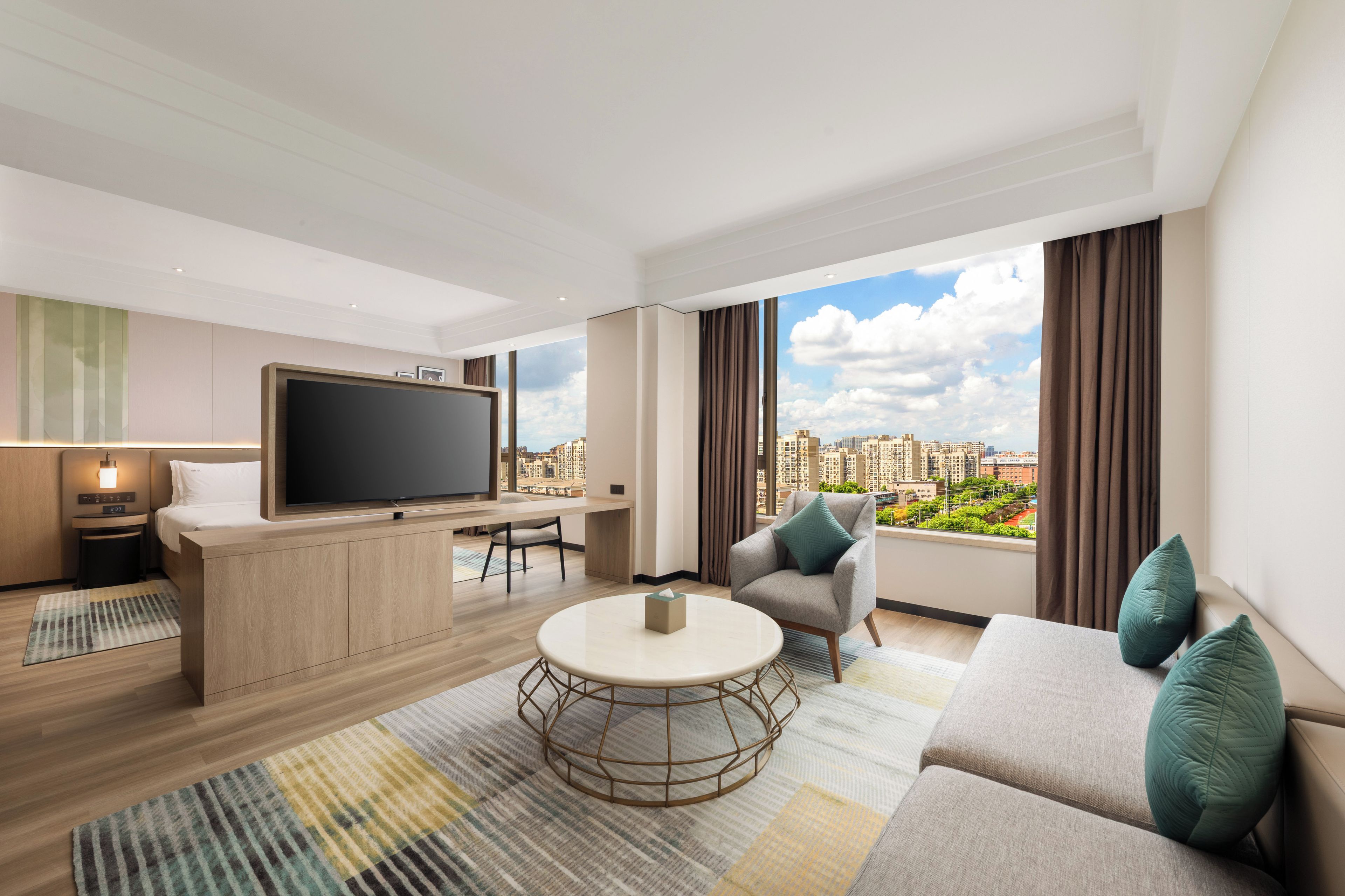 Ein modernes Hotelzimmer mit großem Fernseher, einer Couch, einem Sessel und einem Esstisch.