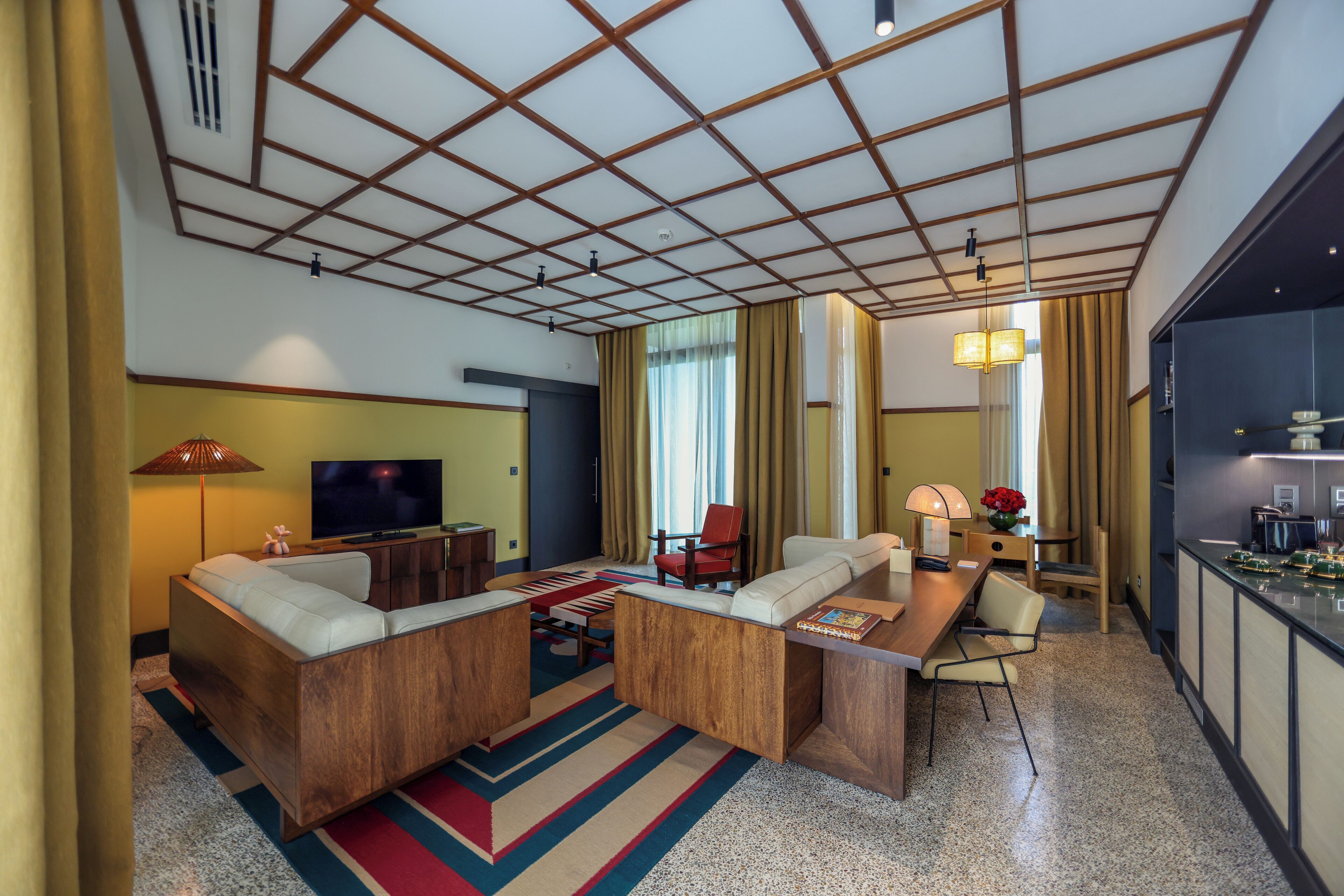 A modern hotel room with a wooden ceiling, a flat-screen TV, a sofa, a dining table, and a kitchen area.
