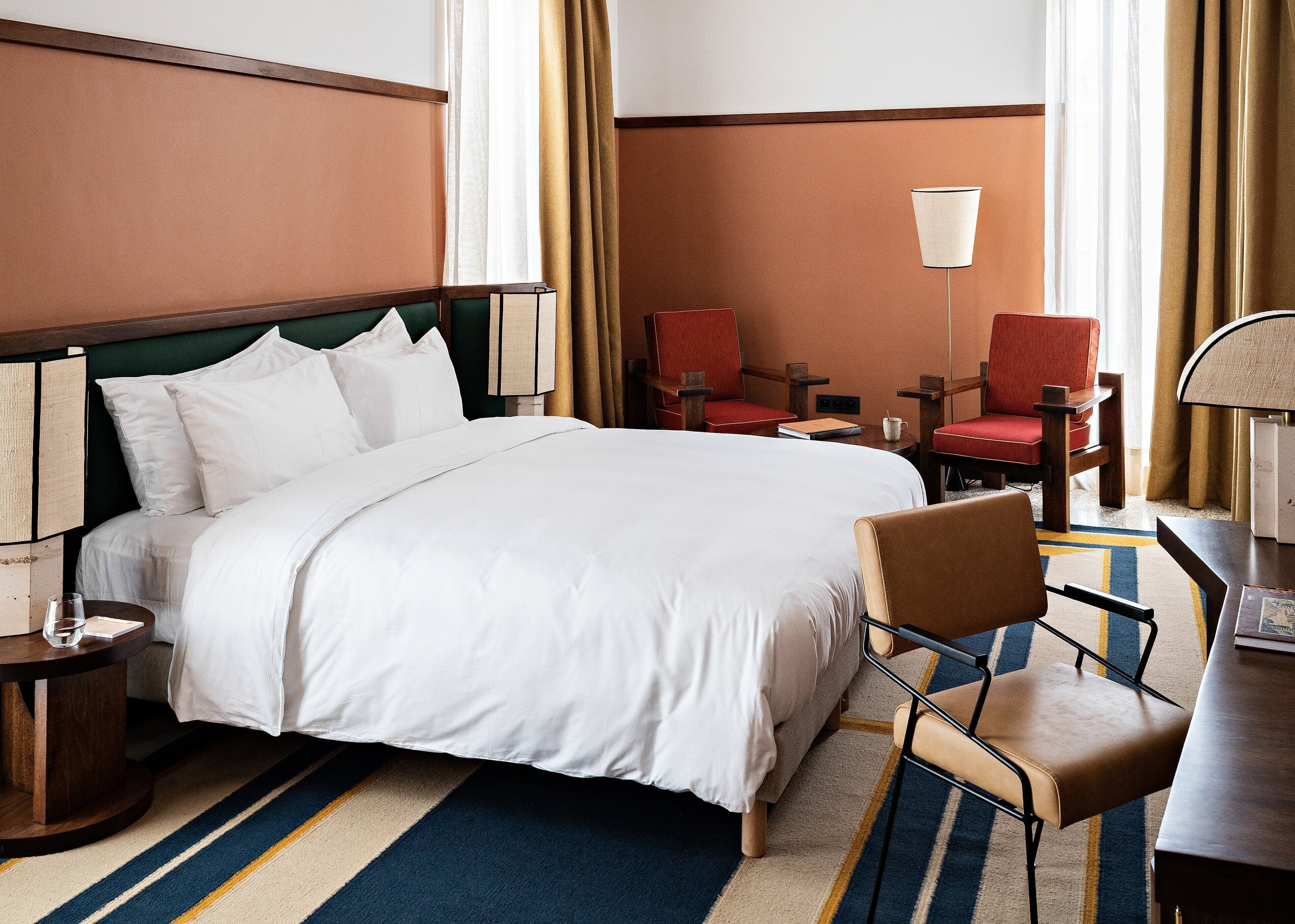 A hotel room with a large bed, two red chairs, a desk with a lamp, and a window with curtains.