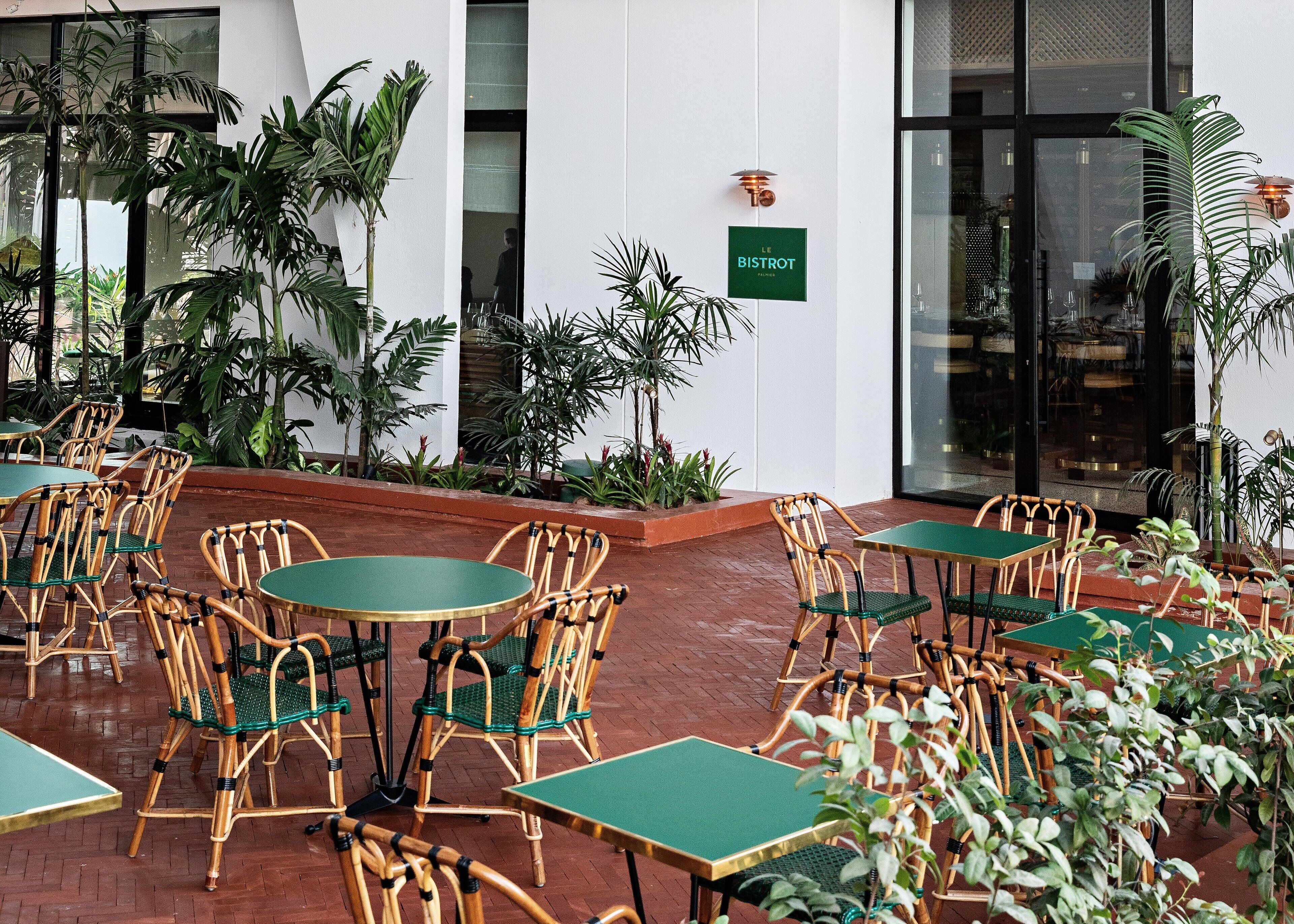 Un área de estar al aire libre con sillas de mimbre y mesas verdes, rodeada de plantas en macetas y un letrero que dice "LE BISTROT".