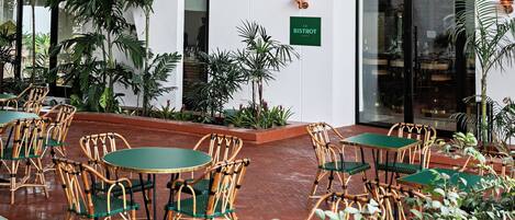 Un área de estar al aire libre con sillas de mimbre y mesas verdes, rodeada de plantas en macetas y un letrero que dice "LE BISTROT".