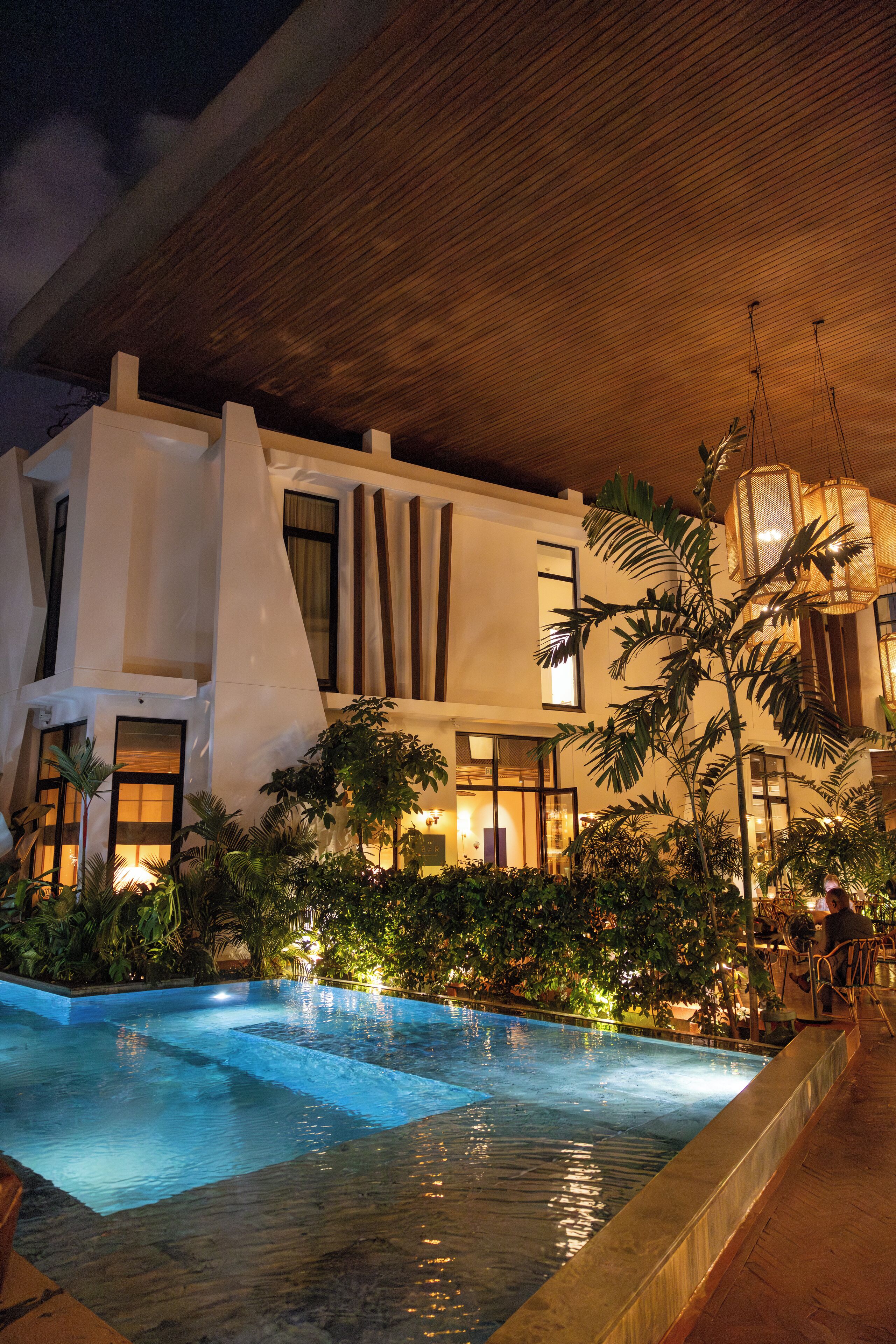 A modern hotel with a swimming pool, lush greenery, and a well-lit interior.
