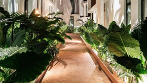 Un pasillo flanqueado por macetas con plantas, que conduce a un edificio con grandes ventanales.