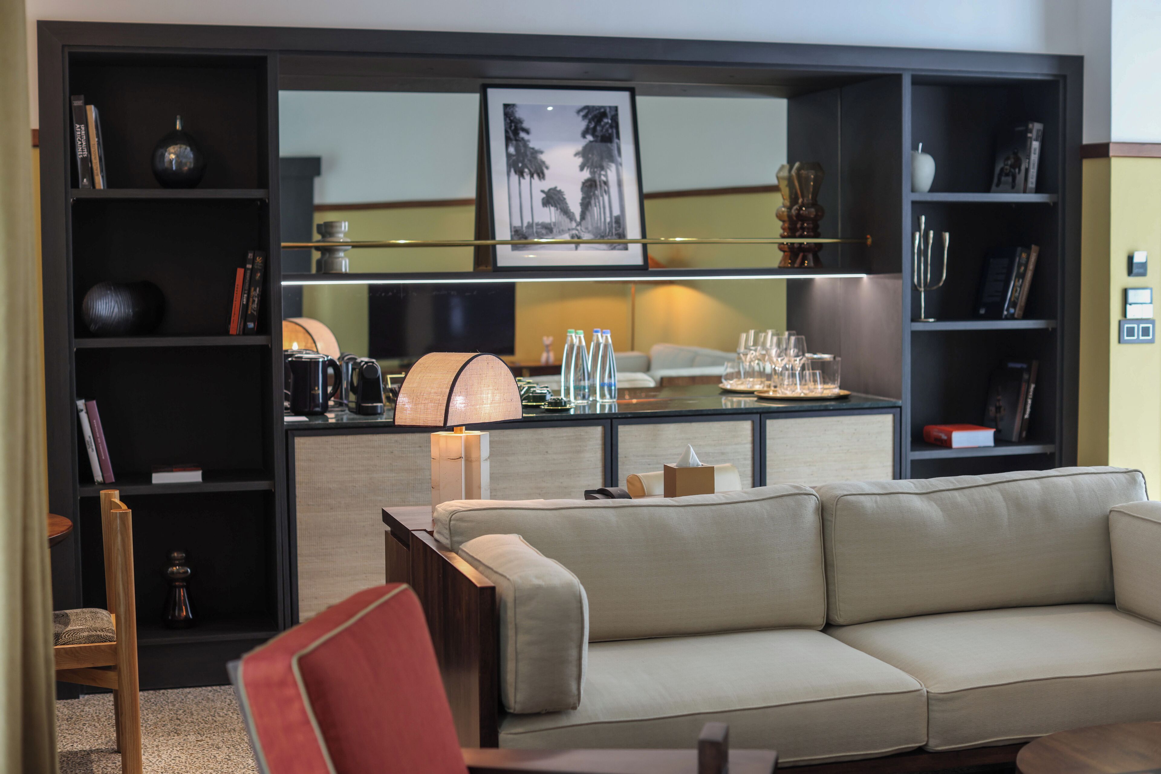 Uma sala de estar moderna com um sofá bege, uma poltrona vermelha e uma mesa de centro. Há estantes com diversos objetos, um abajur e um quadro na parede.
