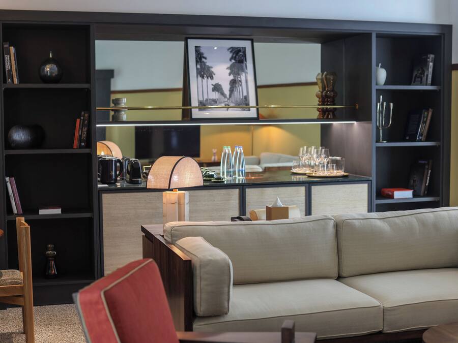 Uma sala de estar moderna com um sofá bege, uma poltrona vermelha e uma mesa de centro. Há estantes com diversos objetos, um abajur e um quadro na parede.