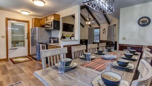 A spacious dining area with a wooden table set for six, surrounded by wooden chairs. A kitchen with modern appliances and a dining area with a chandelier is visible in the background. - Pigeon Forge Getaway, Hot Tub, Game Room, Wifi, $560 FREE tickets each paid day! (Pigeon Forge)