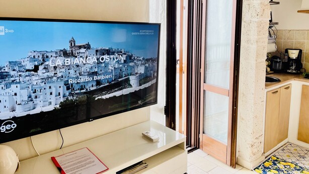 Televisão de tela plana na parede exibindo vista panorâmica de uma cidade costeira, área de cozinha com máquina de café e porta que dá para um espaço ao ar livre.