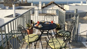 Terraço no terraço com uma mesa posta para dois, exibindo um copo de suco vermelho, um pastel e uma xícara. O terraço possui cadeiras de metal e um cacto em vaso.