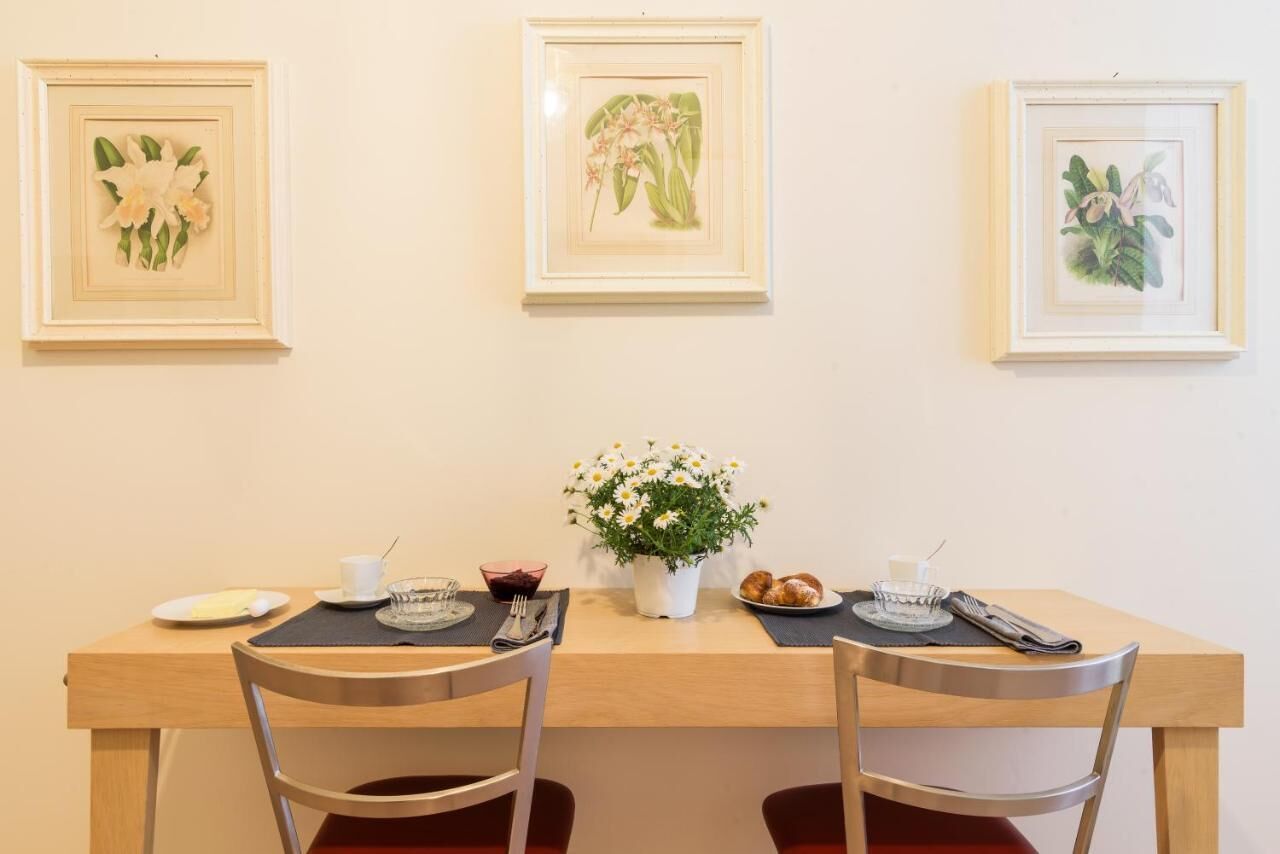 A dining area with a wooden table, two chairs, a vase with white flowers, and framed botanical prints on the wall.