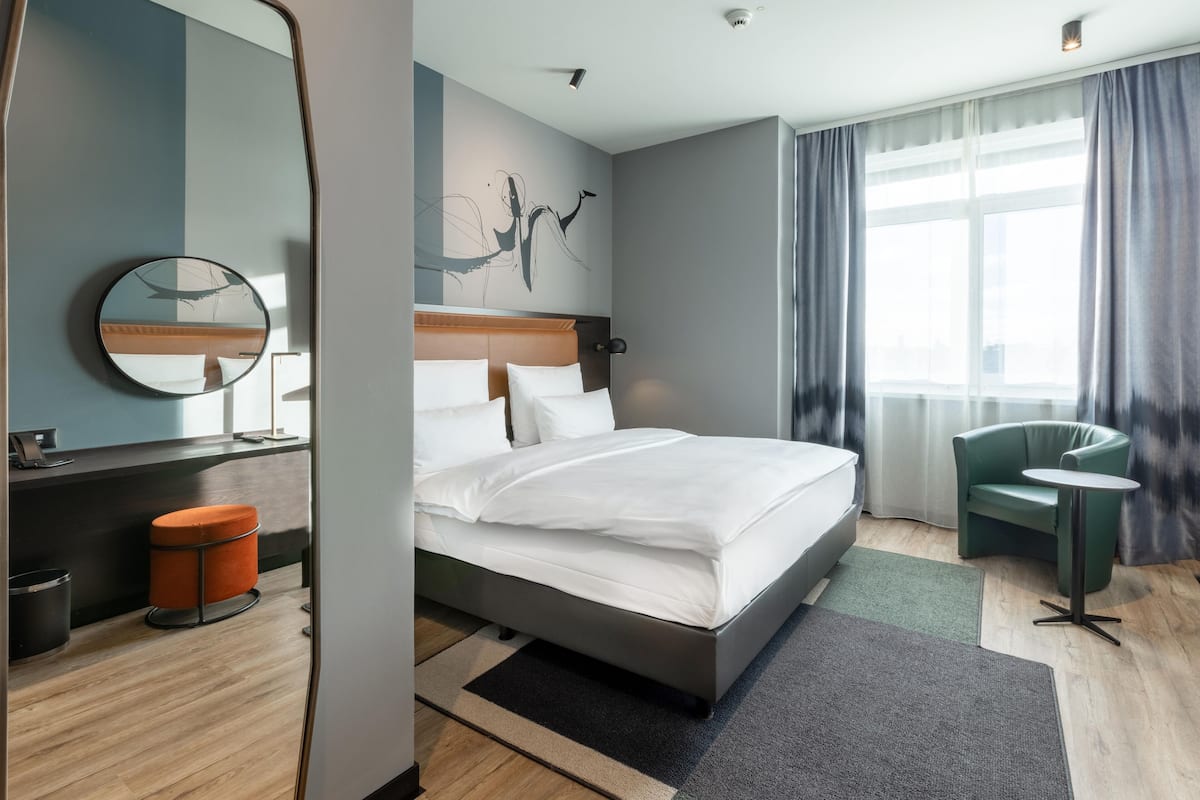 A modern hotel room with a bed, a round mirror, a green armchair, and a small table.