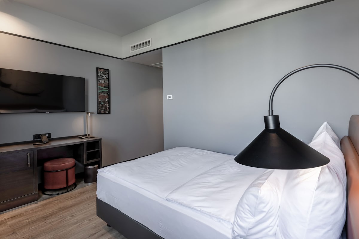 A modern hotel room with a bed, a desk, a chair, a TV, and a lamp.