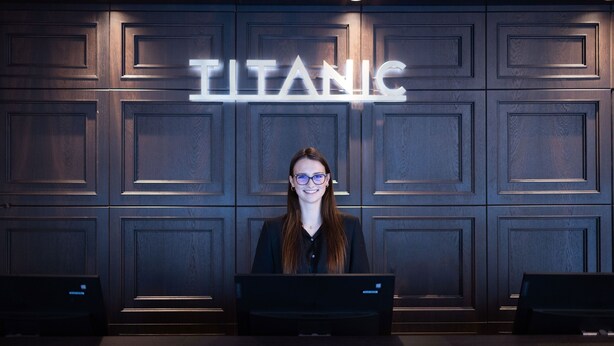 A person seated at a desk with a laptop in front of a wall with a 'TITANIC' sign. - TITANIC Comfort Kurfürstendamm (Berlin)
