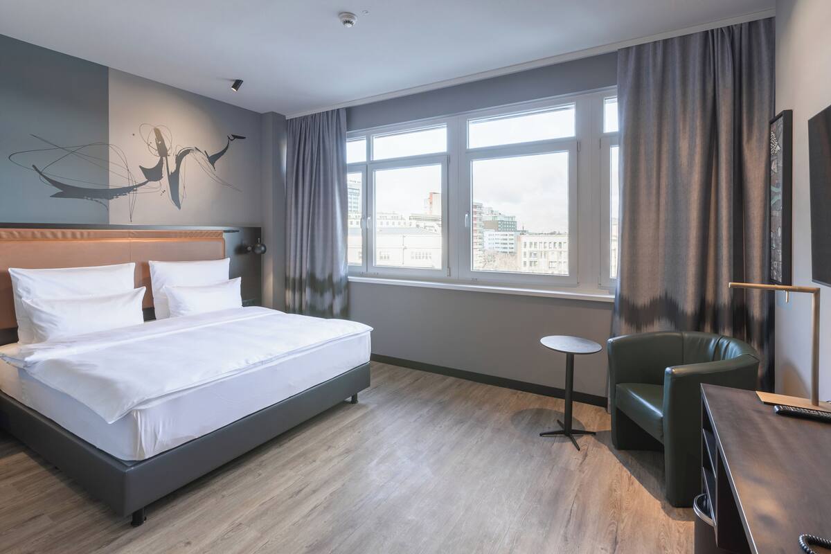 A hotel room with a large bed, a desk, a chair, a small table, and a view of the city through large windows.