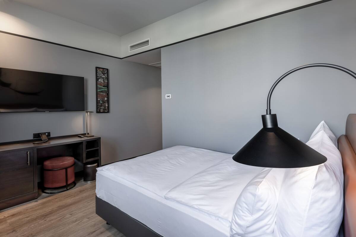 A modern hotel room with a bed, a desk, a chair, a TV, and a lamp.