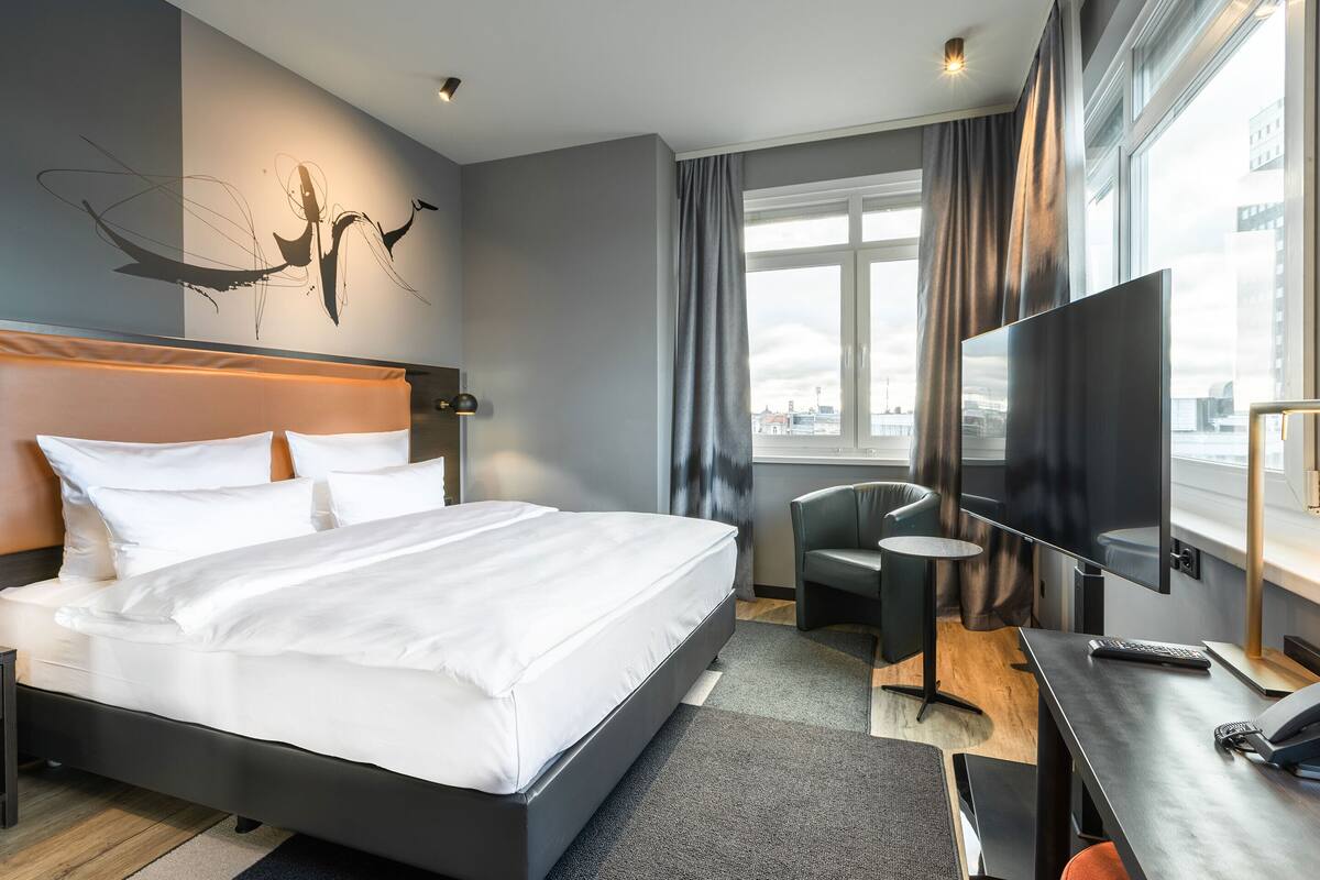 A modern hotel room with a large bed, a desk with a computer, a TV, and a view of the city through large windows.