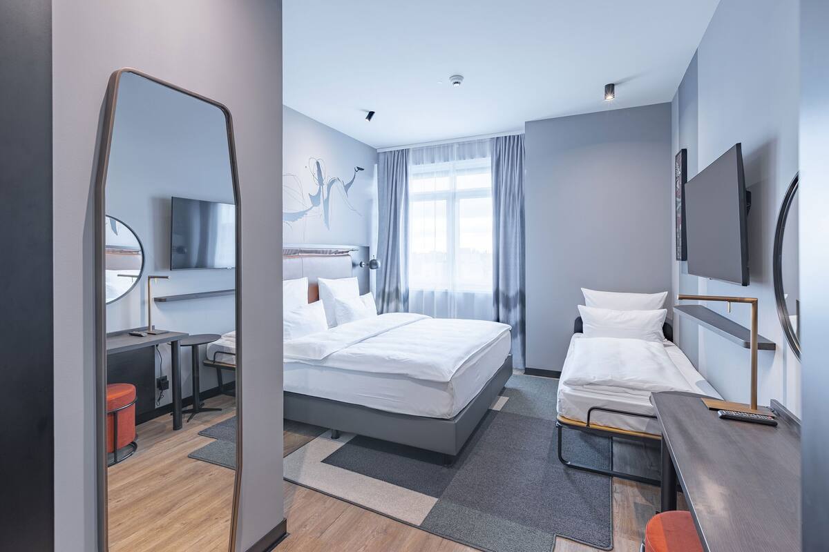 A modern hotel room with a large bed, a desk, a chair, a TV, and a window with curtains.