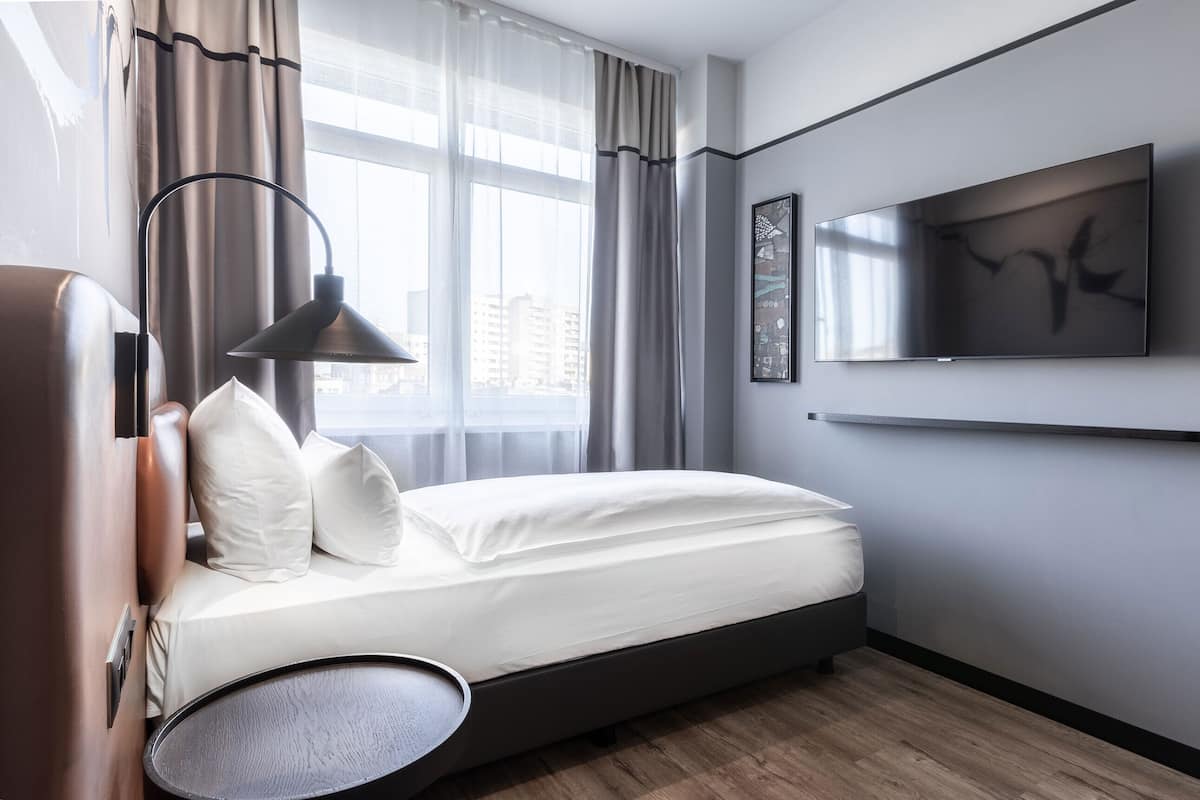 A modern hotel room with a bed, a TV mounted on the wall, and a view of the city through large windows.