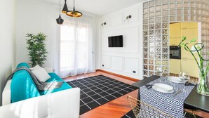 A modern living room with a sofa, a dining table set for two, a large window with sheer curtains, and a wall-mounted TV. - MyHomeInComo - Santa Chiara Apartment (Como)