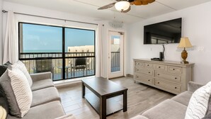 A modern living room with a sofa, a coffee table, a TV, a ceiling fan, and a view of the beach.