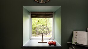 A room with a window, a wall clock, a small table with a decorative tree and tomatoes, and a book on the right.