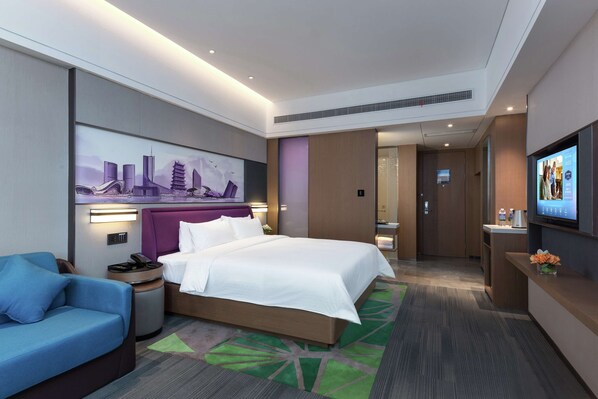 Une chambre d’hôtel moderne avec un grand lit, un canapé bleu, un tableau représentant un paysage urbain, une télévision et un comptoir de réception.
