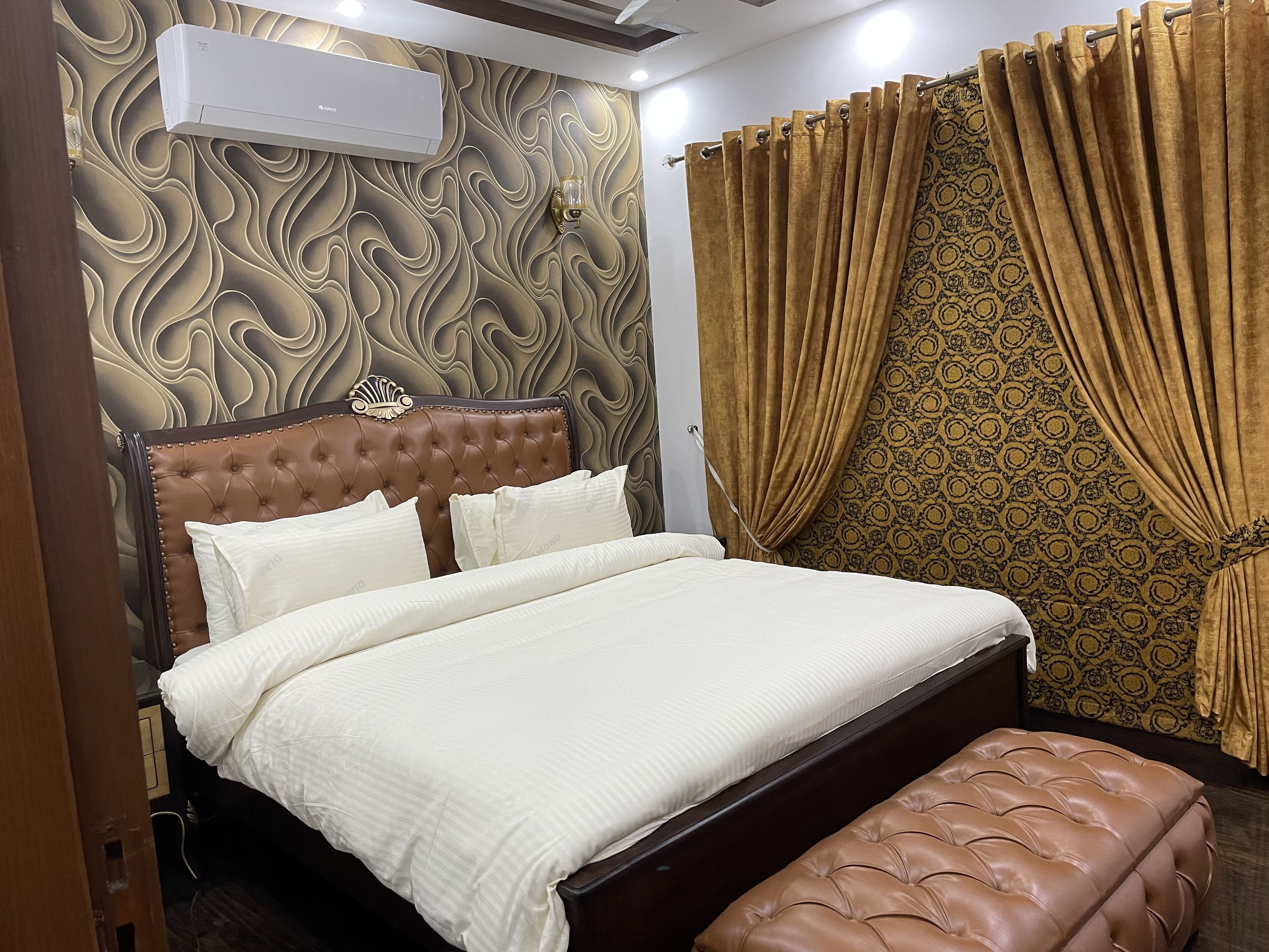 A bedroom with a tufted headboard, a bed with white linens, a brown leather ottoman, and patterned curtains.