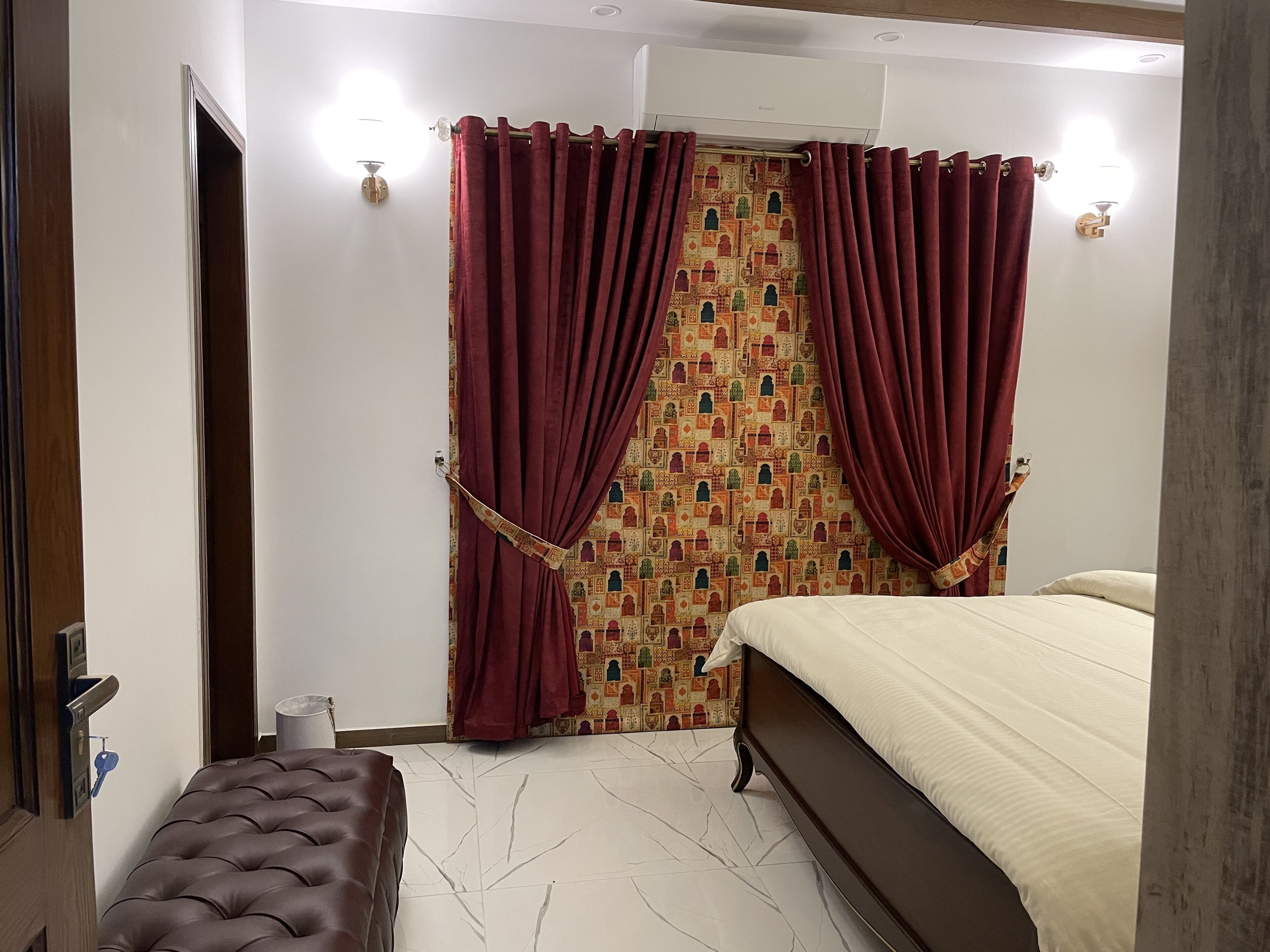 A bedroom with a bed, a bench, a patterned curtain, and a wall-mounted air conditioner.
