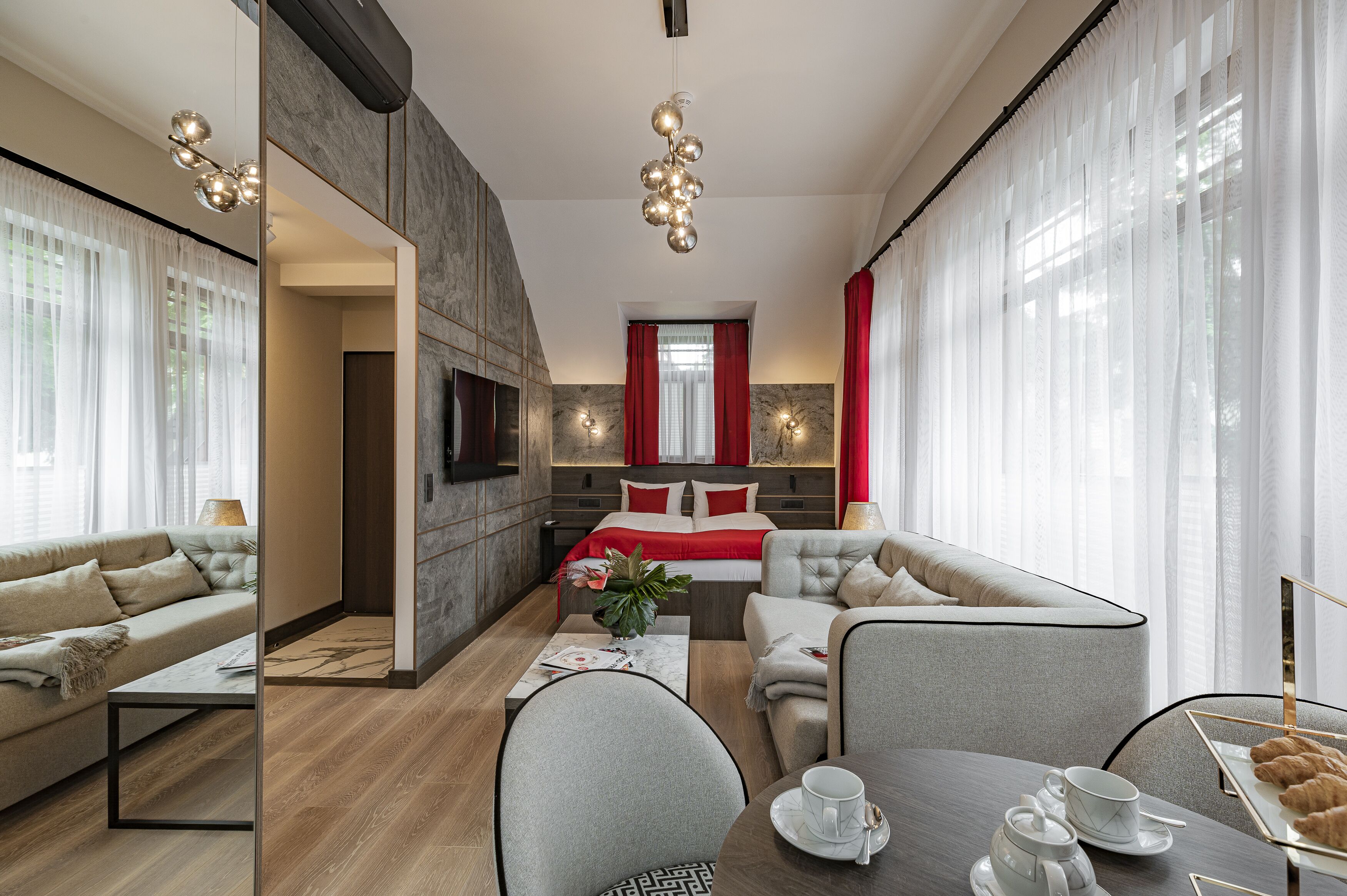 A modern hotel room with a sofa, a dining table with a coffee set, a bed with red bedding, and a TV mounted on the wall.