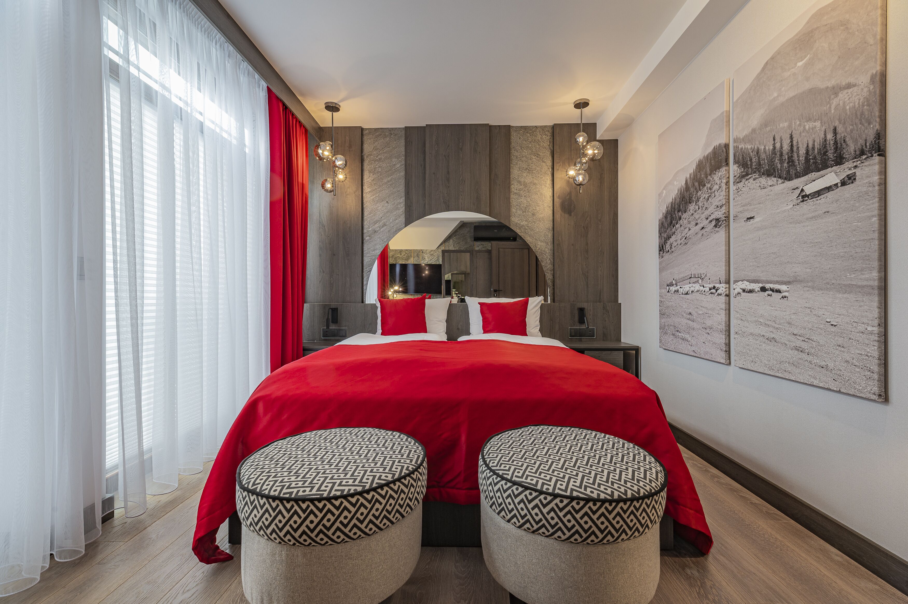 A modern bedroom with a large bed, two ottomans, a mirror, and a mountain landscape painting.