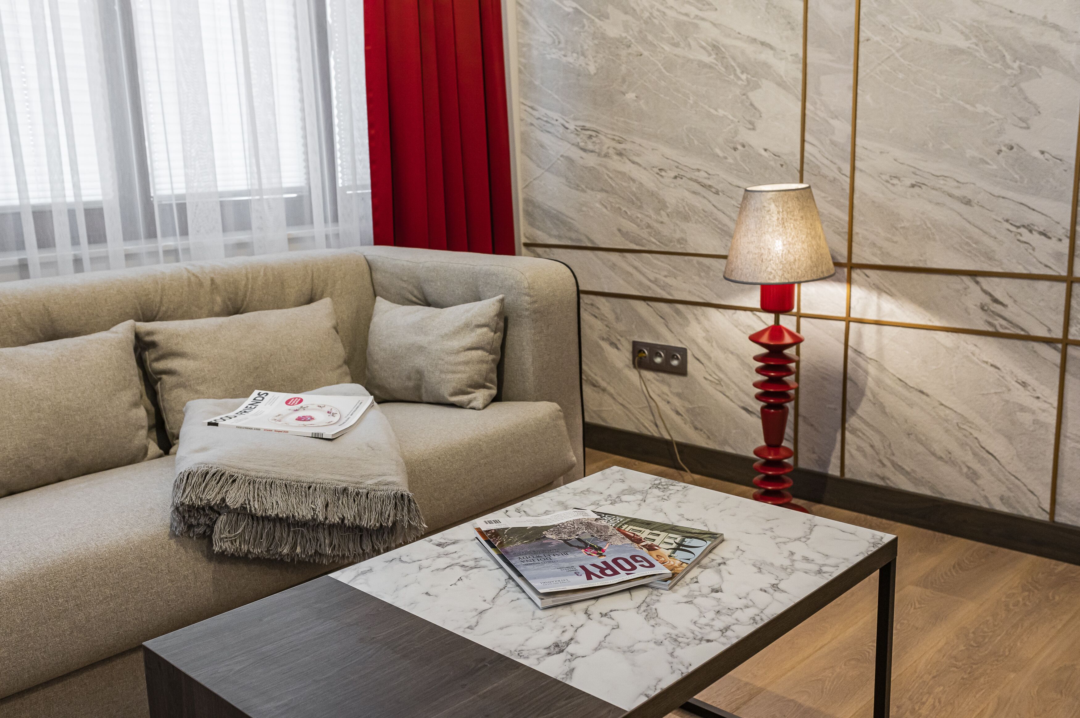 A modern living room with a sofa, a coffee table with magazines, a red lamp, and a marble wall.