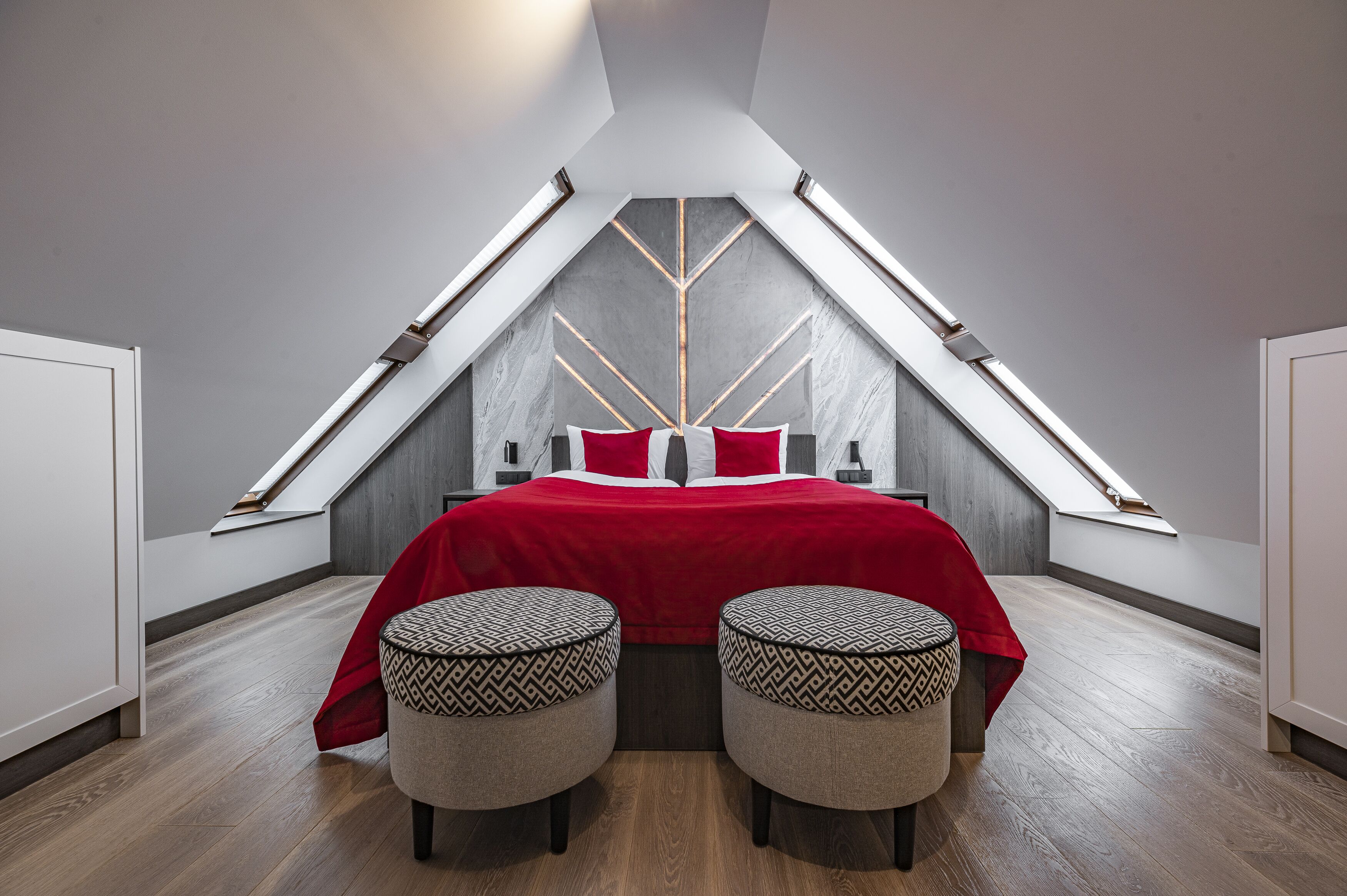 A modern bedroom with a sloped ceiling, a bed with red bedding, two patterned ottomans, and a skylight.