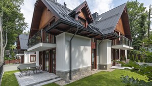 Una casa moderna de dos pisos con techo inclinado, ventanas grandes y un patio con mobiliario exterior.