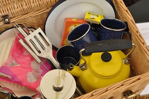 A wicker basket containing a yellow teapot, blue mugs, a spatula, a plate, a box of marshmallows, and a pink bag.