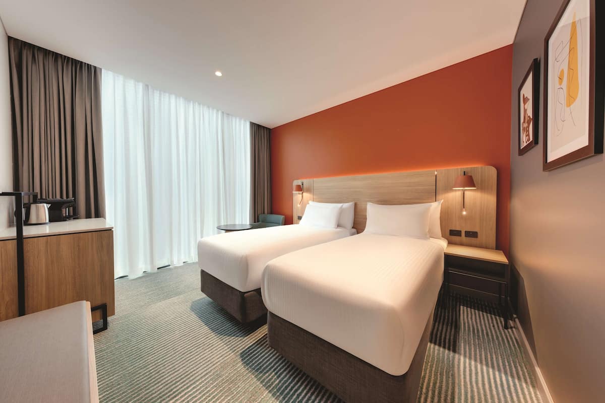 A hotel room with two beds, a desk, a chair, a coffee maker, and a large window with curtains.