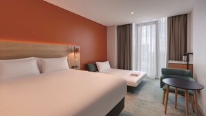 Habitación de hotel con dos camas, una mesita, una silla y vistas a la ciudad a través de amplios ventanales.
