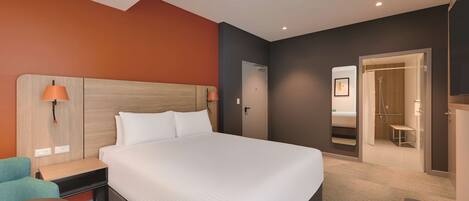 Habitación de hotel con una cama grande, un escritorio, una silla y un baño visible a través de una puerta abierta.