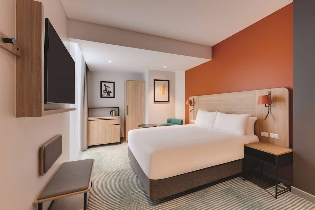 A modern hotel room with a large bed, a desk, a chair, a TV, and a small table with a lamp.