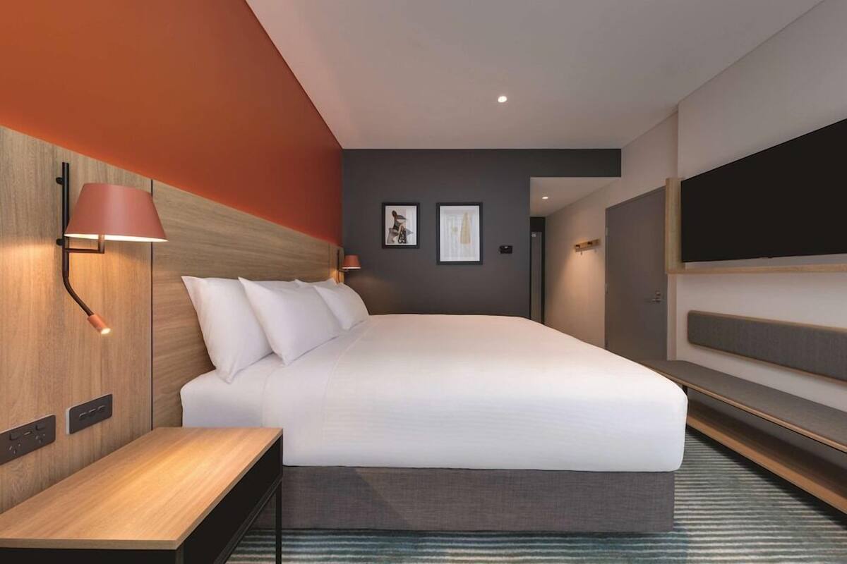 A modern hotel room with a large bed, a bedside table, a wall-mounted TV, and a red accent wall.