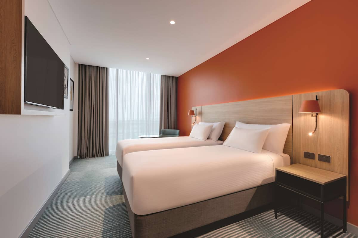 A hotel room with a large bed, a bedside table, a TV mounted on the wall, and a window with curtains.